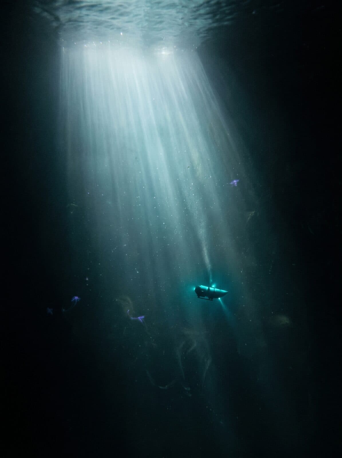 A lone submersible descending through a cathedral shaft of surface light into the deep ocean.